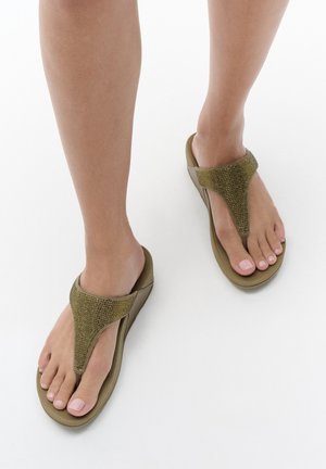 Voeten die olijfgroene teenslippers met gestructureerde bandjes dragen op een witte achtergrond.