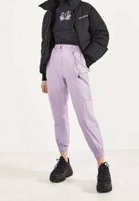 Fioletowe spodnie cargo z elastycznymi ściągaczami mają dwie boczne kieszenie oraz akcent w postaci łańcucha. W zestawie z czarną kurtką puchową i masywnymi czarnymi trampkami.