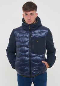 Marineblauwe pufferjack met een gewatteerd ontwerp, voorzien van een ritssluiting op de borst en een capuchon. Gemaakt van glanzend materiaal met contrasterende matte mouwen.