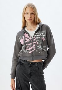 Sudadera corta gris con cremallera frontal, que presenta diseños gráficos y texto en rosa. Tejido suave, mangas largas con puños acanalados, estilo casual.