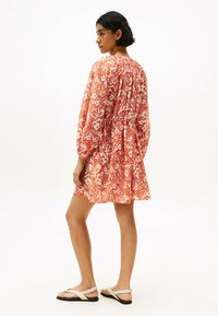 Robe à imprimé floral rouge et blanc avec manches longues bouffantes, légèrement froncée au niveau du col, et coupe ample, associée à des sandales plates.