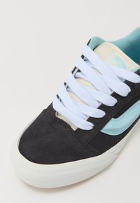 Svarte semskede sneakers med lyseblå detaljer, hvite snører og en kremfarget tunge. Har gummisåle og et strømlinjeformet design.