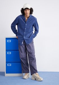 Young person with curly hair wearing white cap, blue button-up shirt, gray cargo pants, and beige sneakers, leaning on blue filing cabinet.