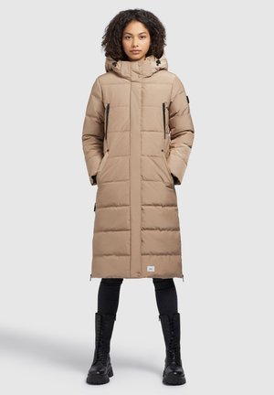 Long, puffy beige coat with a hood, quilted pattern, side pockets with zippers, and a branded patch on the lower hem. Black boots complete the look.