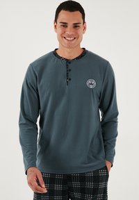 Tums garām, tumši zila vilnas krekls ar pogāmu apkakli un raibu apdari; uz krūtīm ir logotipa plāksne.