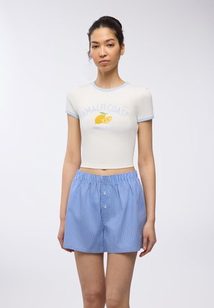Top corto bianco a maniche corte con bordature blu e stampa "AMALFI COAST"; abbinato a pantaloni corti a righe bianche e blu con bottoni.