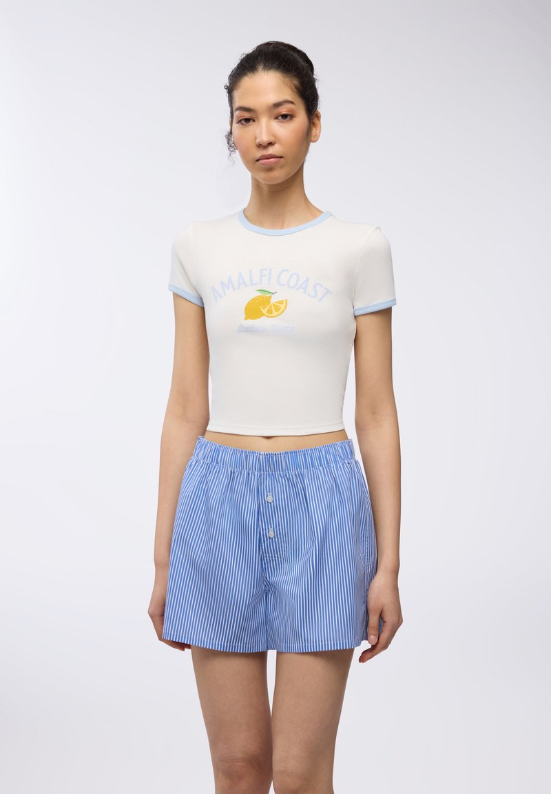 Top corto bianco a maniche corte con bordature blu e stampa "AMALFI COAST"; abbinato a pantaloni corti a righe bianche e blu con bottoni.