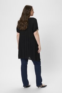 Tunique noire légère, ample, avec des manches courtes retroussées, des fentes latérales et un ourlet arrière long, assortie à un jean bleu foncé et des chaussures noires.
