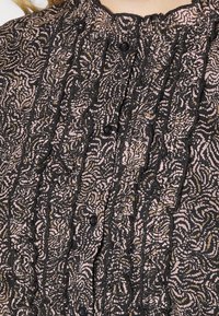 Blouse noire avec un motif floral complexe en rose et or, dotée de volants, de boutons et d'accents en dentelle texturée le long de l'encolure et de l'avant.