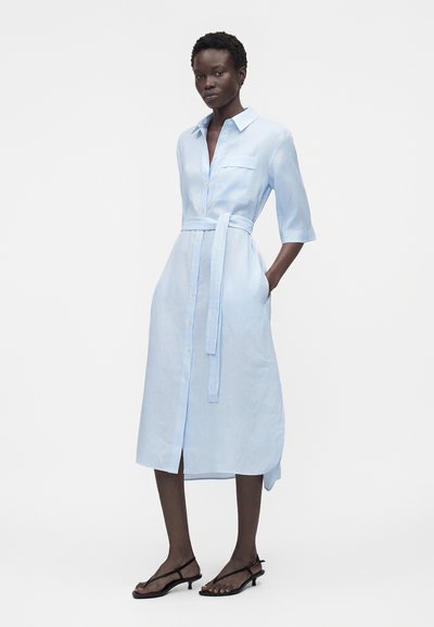 Γυναίκα που στέκεται με ανοιχτό μπλε φόρεμα που κουμπώνει, με ζώνη στη μέση και τσέπες, φορώντας μαύρα σανδάλια με λουράκια και χαμηλό τακούνι.