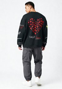 Črna majica s kapuco z rdečo srčno grafiko in dodatnimi risbami na rokavih; v kombinaciji s sivimi cargo hlačami in belimi teniskami.