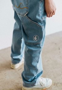 Hellblaue Denim-Jeans mit einer lockeren Passform, die ein einzigartiges gesticktes Design auf der Hintertasche und ein rundes Logodesign aufweisen.