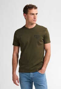 Olivgrünes Baumwoll-T-Shirt mit rundem Halsausschnitt, kurzen Ärmeln und einem blauen Drucklogo auf der linken Brust. Kombiniert mit hellblauen Jeans.