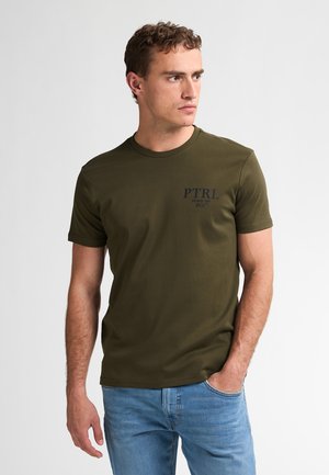 Oliven grøn bomulds T-shirt med rund halsudskæring, korte ærmer og et blåt trykt logo på venstre bryst. Sammen med lyseblå jeans.