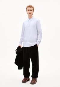 Lichtblauw overhemd met knopen, lange mouwen, gladde stof; zwarte corduroy broek, ontspannen pasvorm; bruine sneakers met accenten; vasthoudend aan een zwart jack.