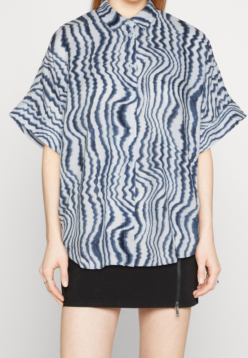 Personne portant une chemise ample à manches courtes avec des rayures ondulées bleues et blanches et une mini-jupe noire avec une fermeture éclair sur le devant.