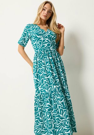Kvinde iført en teal og hvid maxikjole med kortærmet løvprint, stående med den ene hånd på hoften og den anden ved håret.