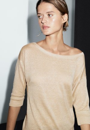 Jeune femme aux cheveux courts portant un haut beige à épaules dénudées, regardant vers la gauche sur un fond divisé en noir et blanc.