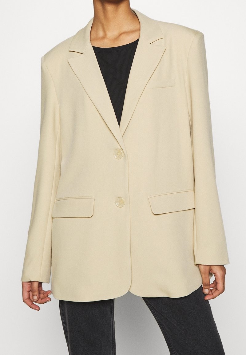 Personne portant un blazer beige à deux boutons avec des poches à rabat sur un haut noir et un pantalon foncé, cadrée sous le cou.