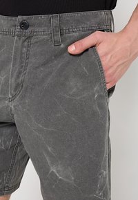 Shorts en denim gris avec un motif marbré subtil, fermeture à bouton, poche et une finition douce au toucher. Gros plan avec une main dans la poche.