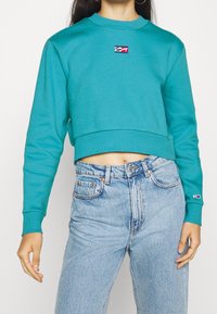 Avskuren turkos sweatshirt i mjukt tyg, med rund halsringning och en logotyp på bröstet, tillsammans med ljusblå jeans.