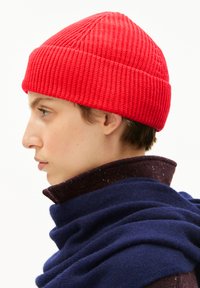 Rode gebreide beanie met een geribbelde textuur en een spitse top, gedragen met een donkere sjaal en een bordeauxrode gekraagde shirt, zijaanzicht.