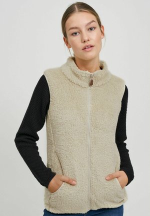 Beige fleecevest med høy krage og glidelås, svarte teksturerte langermede ermer, sidelommer og myk, fluffy tekstur.