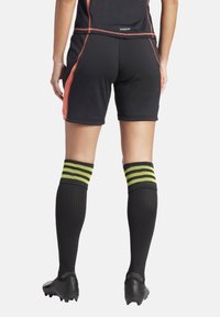 Zwarte sport shorts met oranje stiksels, gecombineerd met kniehoge zwarte sokken met gele strepen. Zwarte voetbalschoenen zichtbaar.