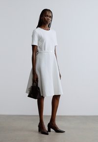 Robe blanche à manches courtes avec un motif texturé, taille cintrée et jupe évasée. Associée à un petit sac à main noir et des talons pointus.