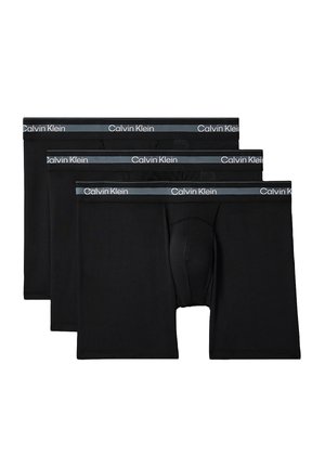 Mężczyzna bez koszulki o muskularnej budowie ciała, ubrany w czarne bokserki Calvin Klein, stojący boso na jednolitym, szarym tle ze skrzyżowanymi rękami.