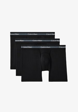 Oberkörperfreier Mann mit muskulösem Körperbau, der schwarze Calvin Klein Boxershorts trägt, barfuß vor einem schlichten grauen Hintergrund stehend mit verschränkten Armen.