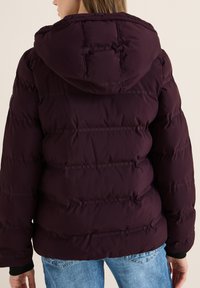 Giacca a piuma bordeaux con cappuccio, caratterizzata da un design trapuntato e polsini elasticizzati, indossata su jeans blu, che mostrano una superficie morbida e testurizzata.