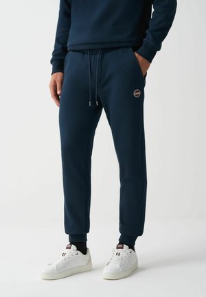Mörkblå sweatpants med dragsko i midjan, ribbade muddar och en rund logotyppatch på fickan. Stylad med vita sneakers.