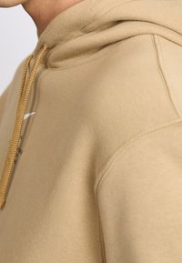 Beige mikina s kapucí vyrobená z měkkého materiálu, s tkaničkou se zlatými akcenty a bílým logem na hrudi.