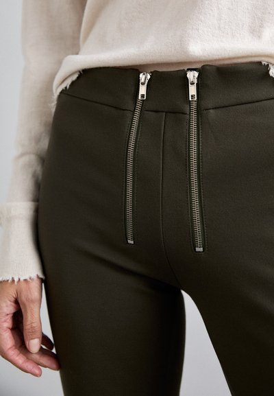 Olijfgroene broek met een hoge taille en twee zilveren ritsaccenten aan de voorkant. De stof lijkt glad met fijne details langs de naden.