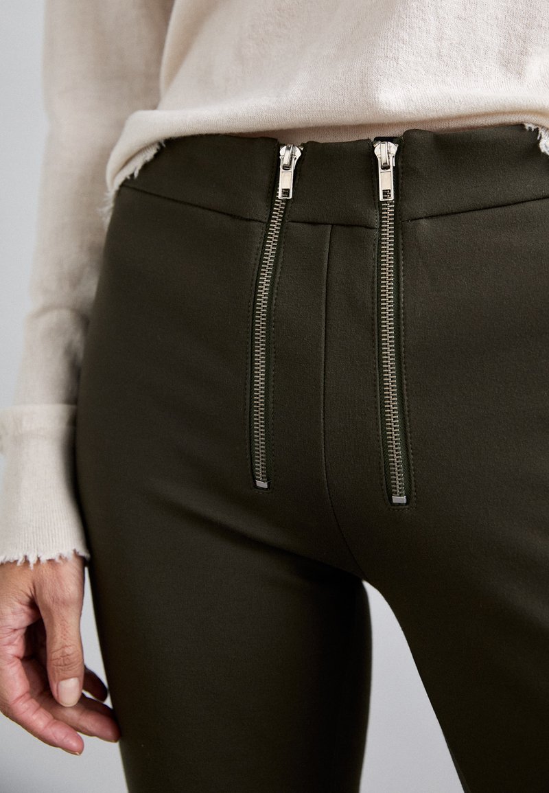 Olive green pants with a high waist and two silver zipper accents at the front. Fabric appears smooth with fine detailing along the seams.