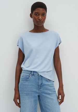 Vrouw staat tegen een witte muur, draagt een lichtblauw T-shirt met korte mouwen in een hoge taille jeans en kijkt recht vooruit met een neutrale uitdrukking.