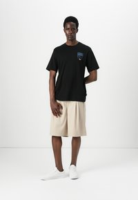 Black cotton T-shirt with a blue graphic logo on the chest, paired with beige pleated shorts and white sneakers. Model stands against a white background.