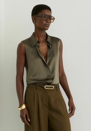 Vrouw draagt een mouwloze olijfgroene satijnen blouse in een hoge bruine broek gestopt met een gouden riem, grote bril en gouden armband.