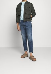 Man med mörkgrön jacka, ljusblå skjorta, rullade blå jeans och beige sandaler som står mot en enfärgad bakgrund.