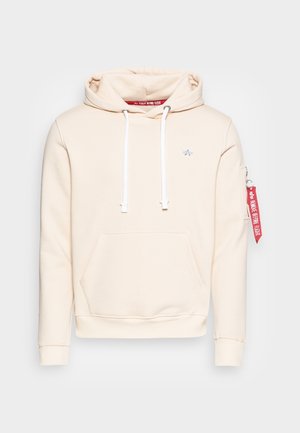 Beżowa bluza z kapturem z kieszenią z przodu, białymi ściągaczami, małym logo na lewej piersi oraz czerwoną zawieszką na lewym rękawie z napisem "Remove Before Flight".