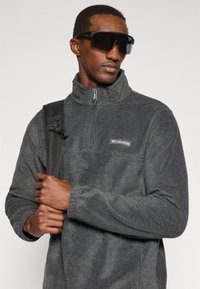 Γκρι φλίς πόλο με ψηλό γιακά και μισό φερμουάρ, που διαθέτει ένα μικρό λογότυπο Columbia, και φοριέται με μια μαύρη τσάντα πλάτης και μεγάλα γυαλιά ηλίου.