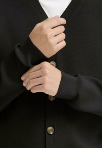 Juodas kardiganas su V formos iškirpte, turintis tris apvalius šviesiai rudos spalvos mygtukus. Raukšlėti rankogaliai ir tekstūruotas audinys.