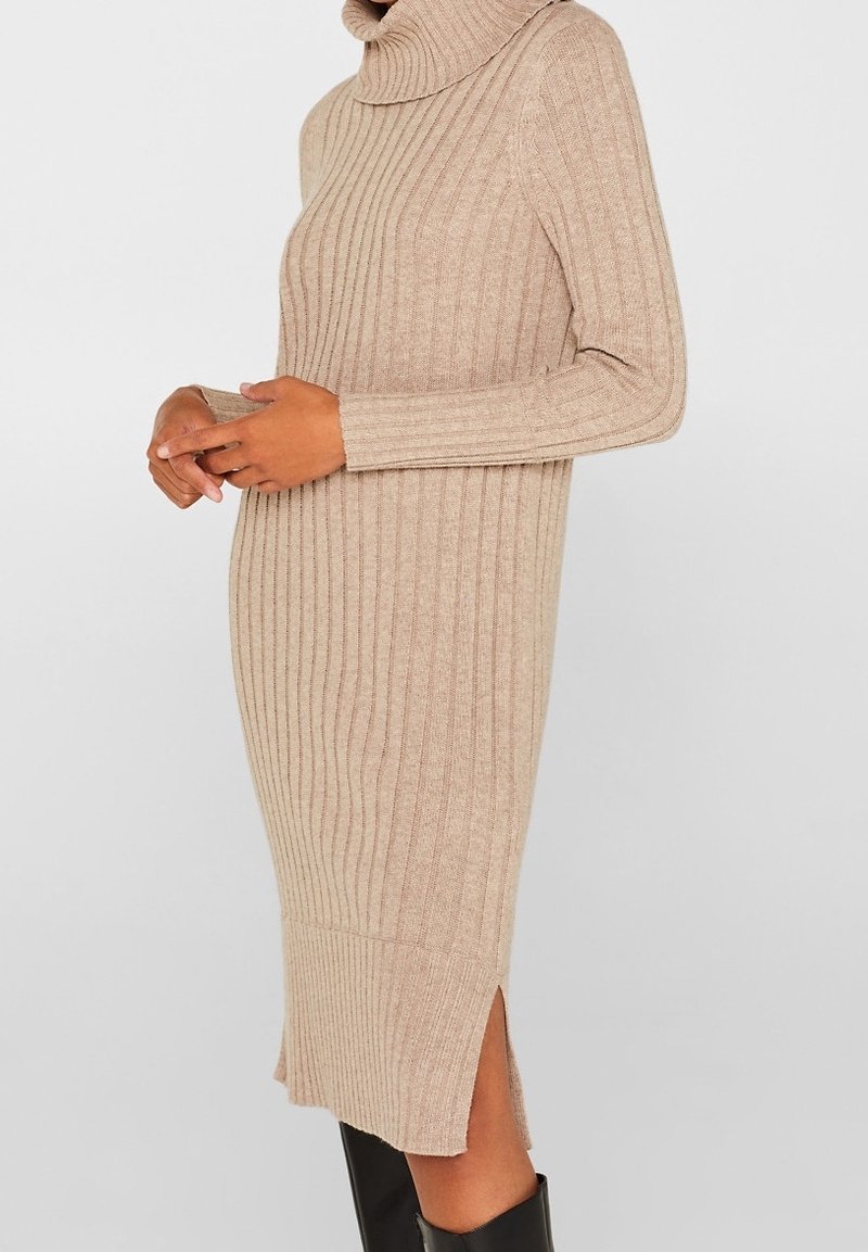 Robe-pull à col roulé en tricot côtelé beige avec fente latérale portée par une personne aux mains croisées et aux bottes noires.