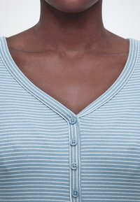 Prikaz izbliza gornjeg dijela prsa i vrata osobe koja nosi plavu i bijelu prugastu košulju s dugmićima i V-izrezom ovratnika.