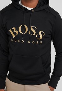 Musta huppari, joka on valmistettu puuvillasekoitteesta. Siinä on suuri kultainen "BOSS"-logo ja kengurun tasku, sekä kiristysnauhat ja resorit hihansuissa.