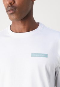 Tricou alb din bumbac, cu un eticheta rectangulară de culoare albastră deschisă, pe care este inscripționat textul "Calvin Klein" în partea stângă sus a pieptului. Guler rotund.