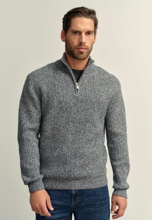 Suéter gris de punto acanalado con cuello con cremallera, que presenta mangas largas y un acabado texturizado, llevado sobre una camiseta negra y jeans azules.