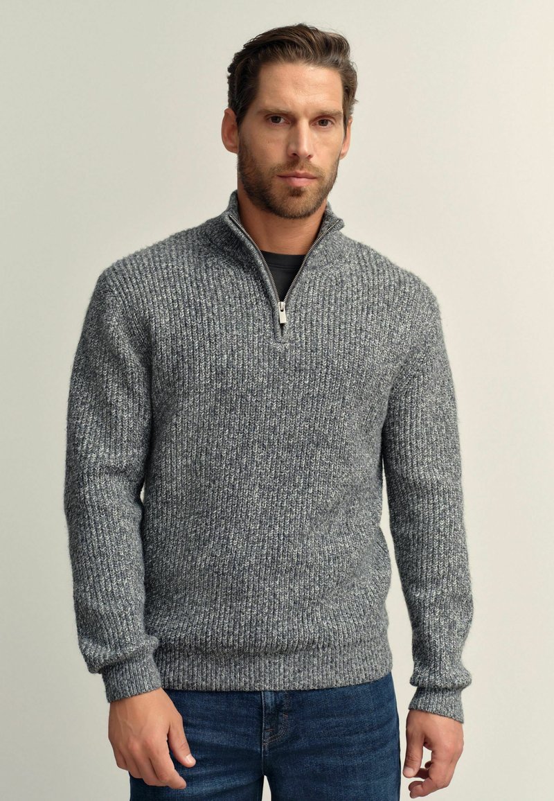 Grauer, gerippter Strickpullover mit Reißverschlusskragen, ausgestattet mit langen Ärmeln und einer strukturierten Oberfläche, getragen über einem schwarzen Oberteil und blauen Jeans.