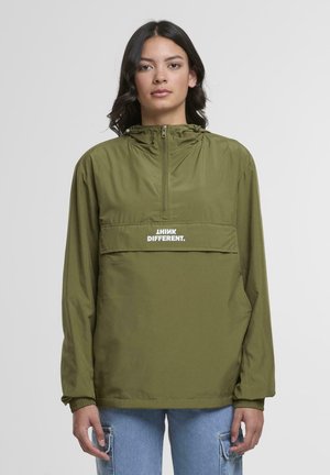 Jeune femme de face portant une veste à capuche olive avec le texte "PENSEZ DIFFÉREMMENT" et un jean bleu clair avec des poches latérales.